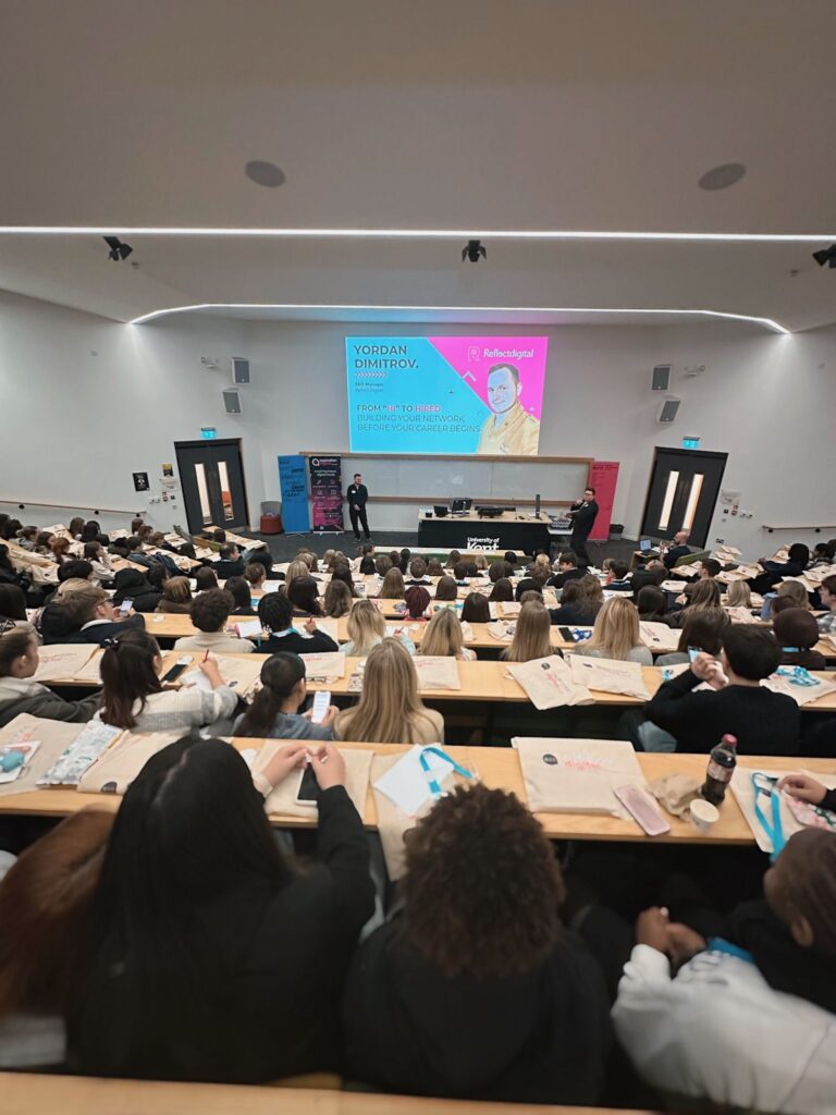 Rows of students in a lecture hall, seen from the back, facing a presentation screen