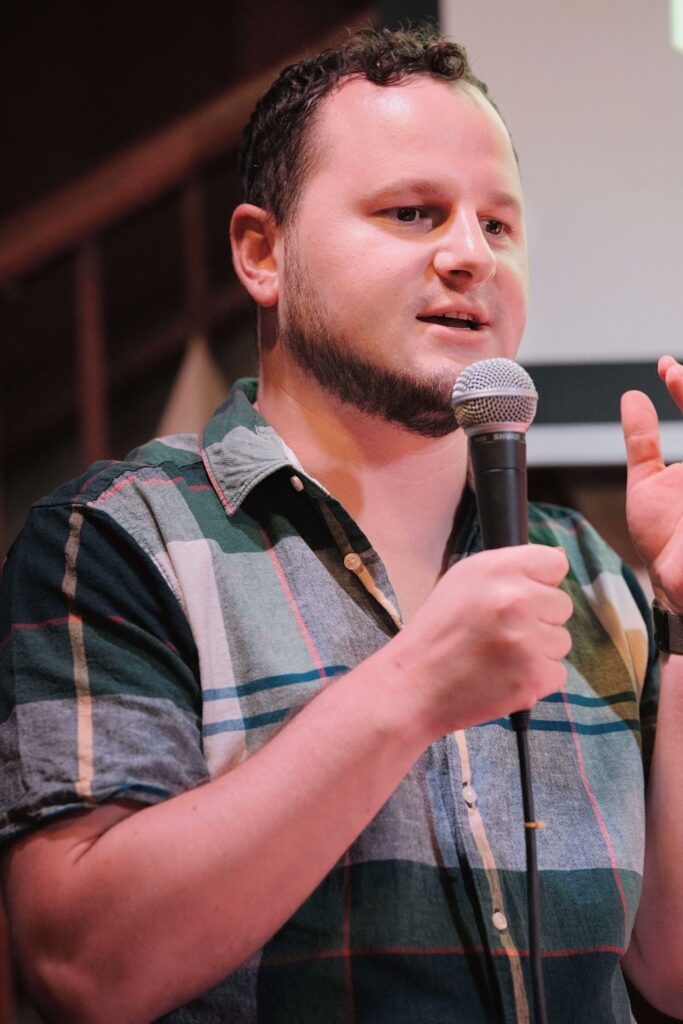 Yordan Dimitrov in a plaid shirt speaking into a handheld microphone and gesturing with one hand